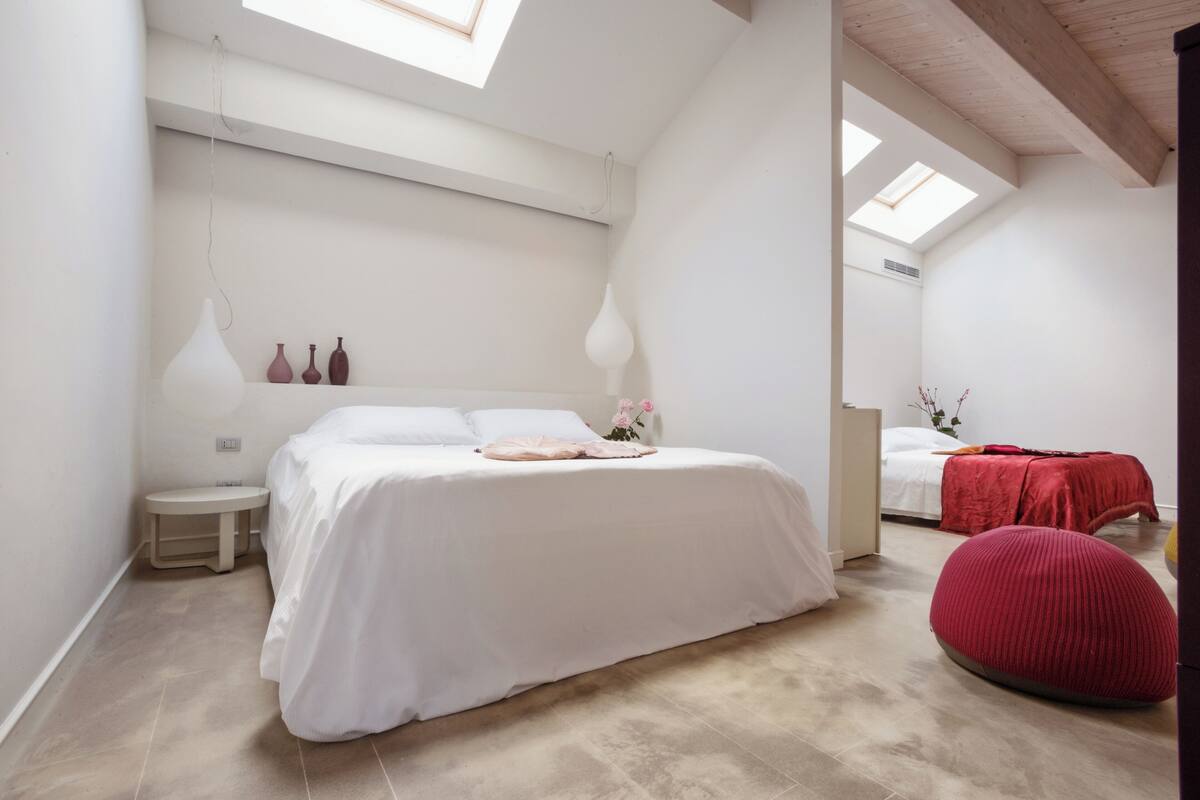 A bedroom with a large bed, a small bedside table, a red bean bag, and a skylight.