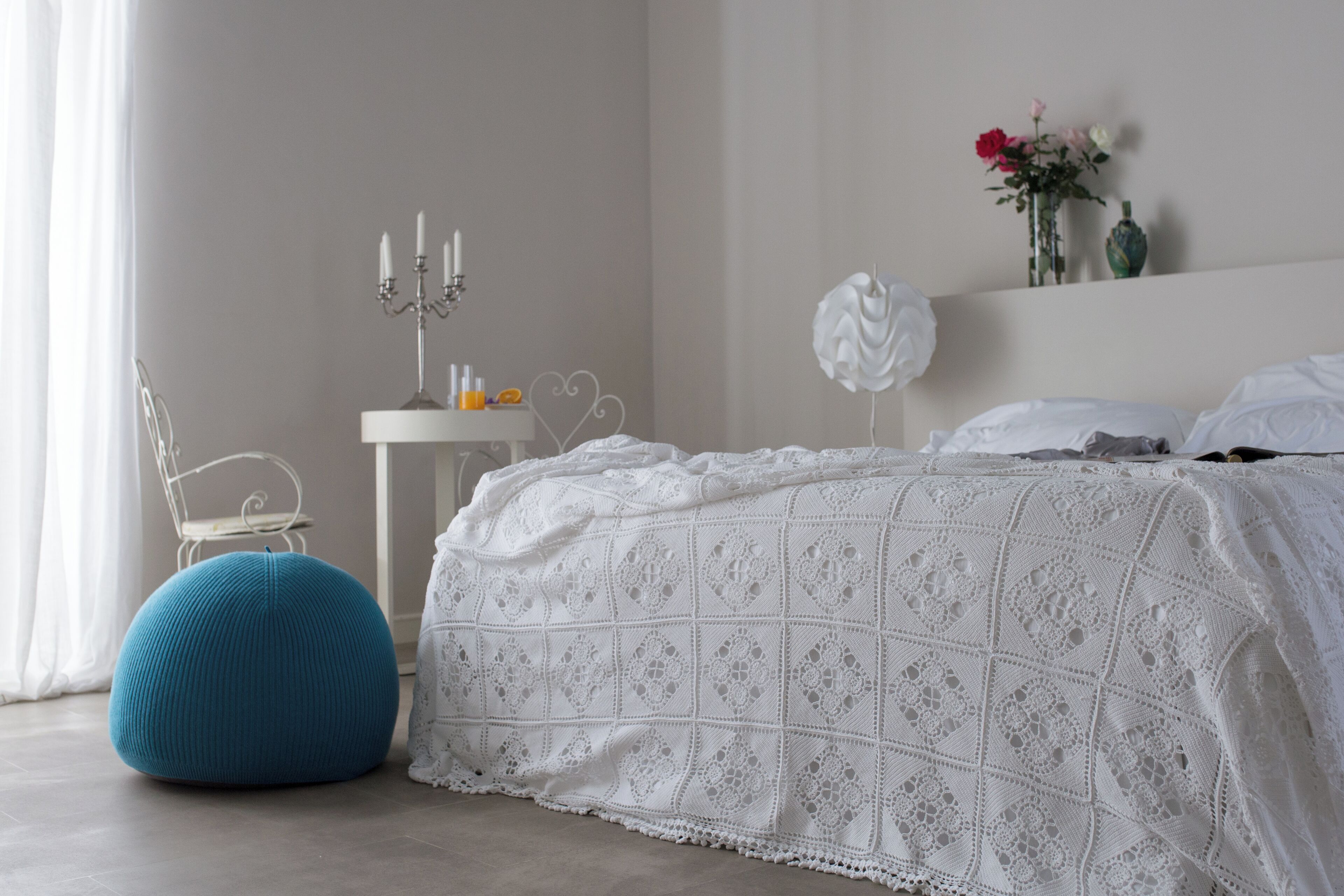 Un dormitorio con una cama, una mesita de noche blanca, un otoman azul, una lámpara blanca y un florero con flores en una estantería.
