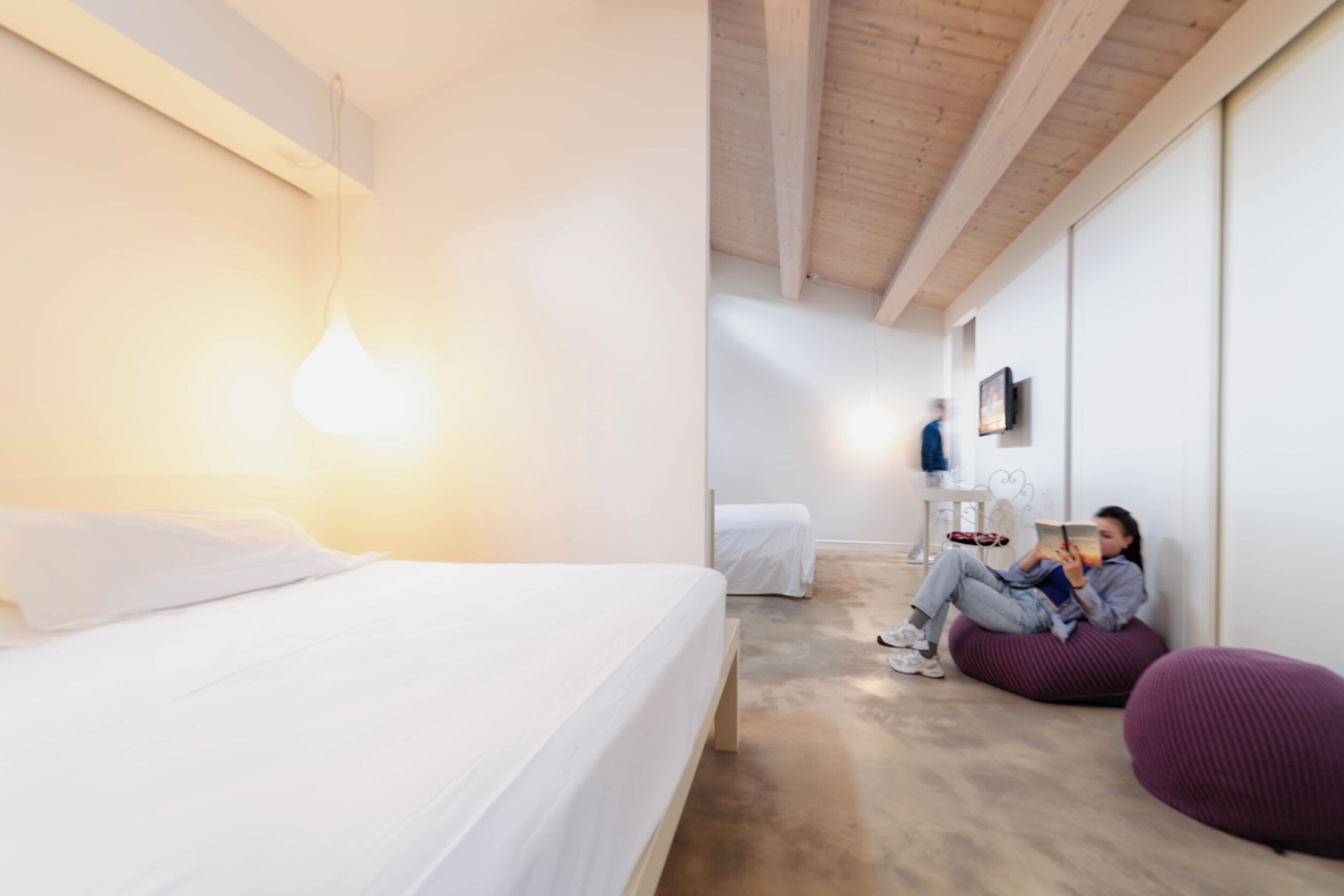 Una habitación de hotel moderna con una cama, un rincón de lectura con sillones de peluche, y un televisor en la pared.
