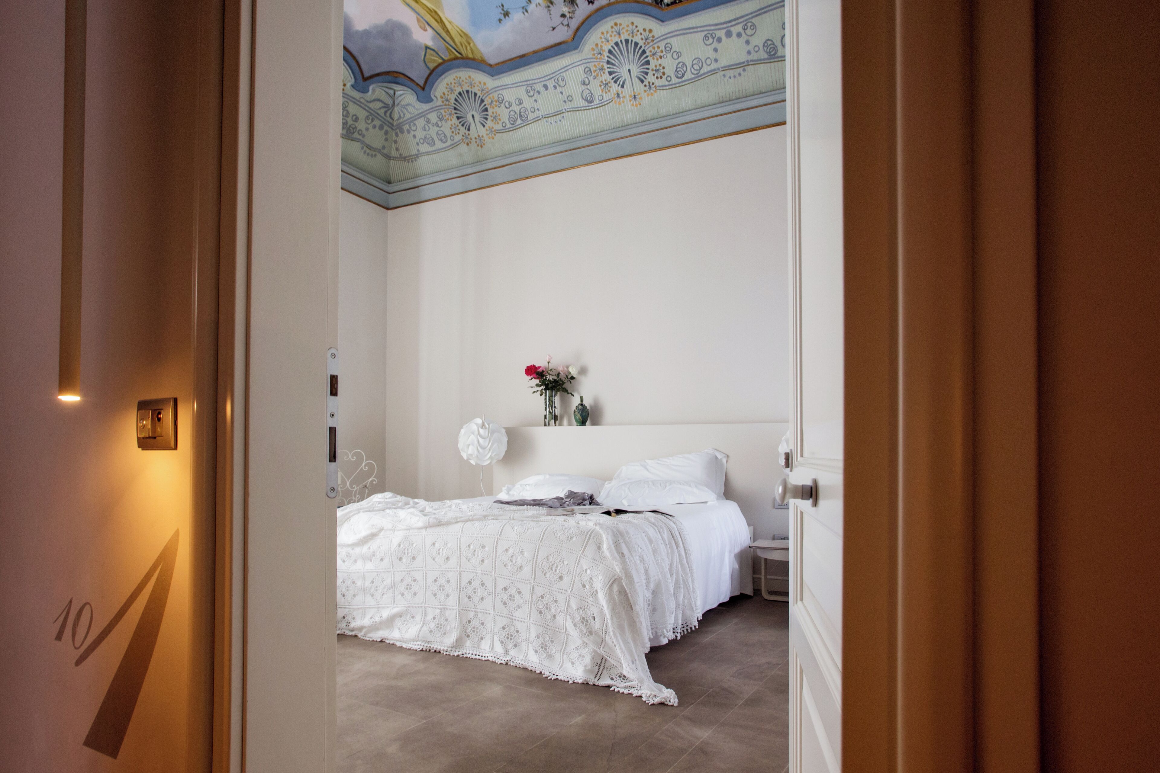 Habitación de hotel con cama, mesita de noche, un florero con flores y un techo con diseño decorativo.
