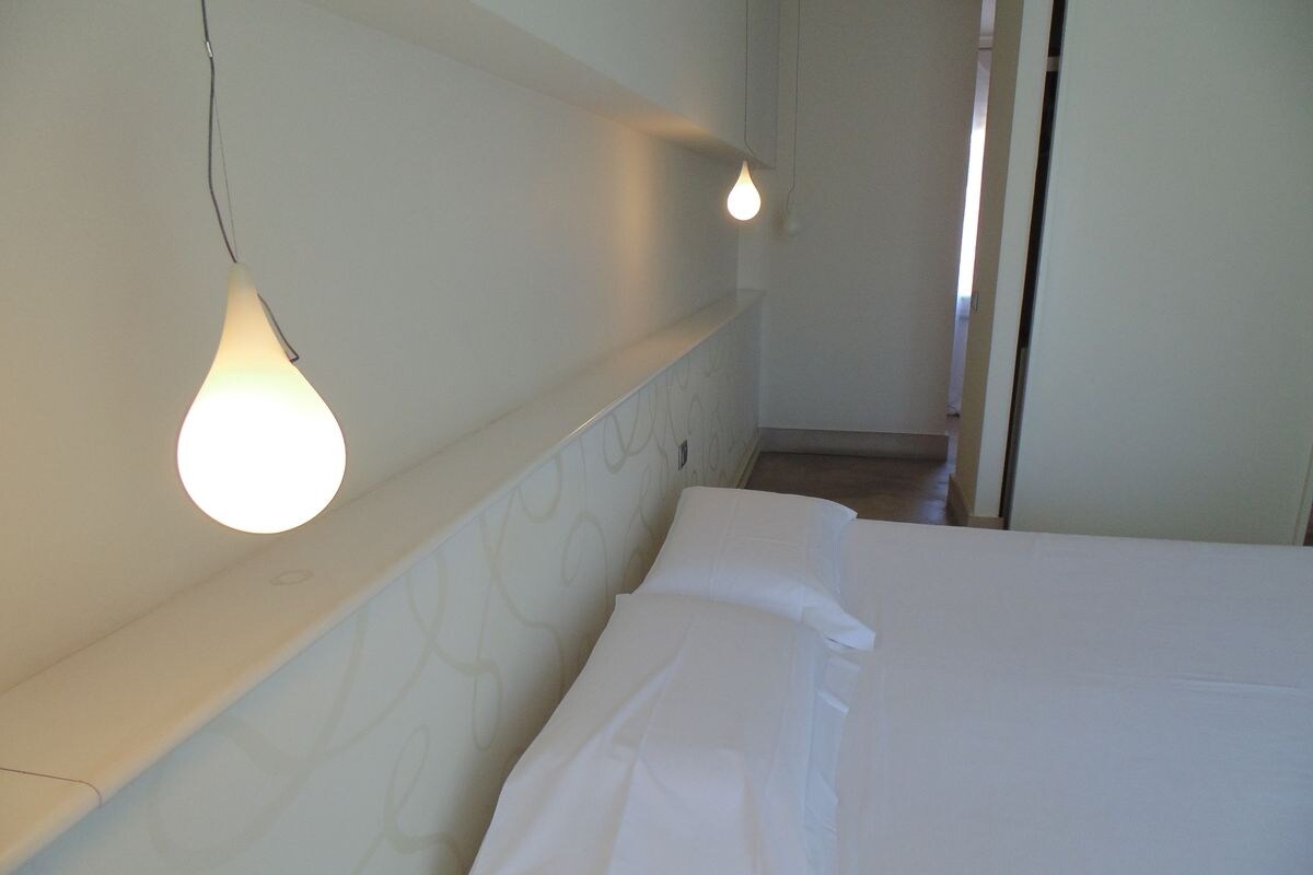 A hotel room with a bed, white bedding, a headboard with a light fixture, and a door leading to another room.