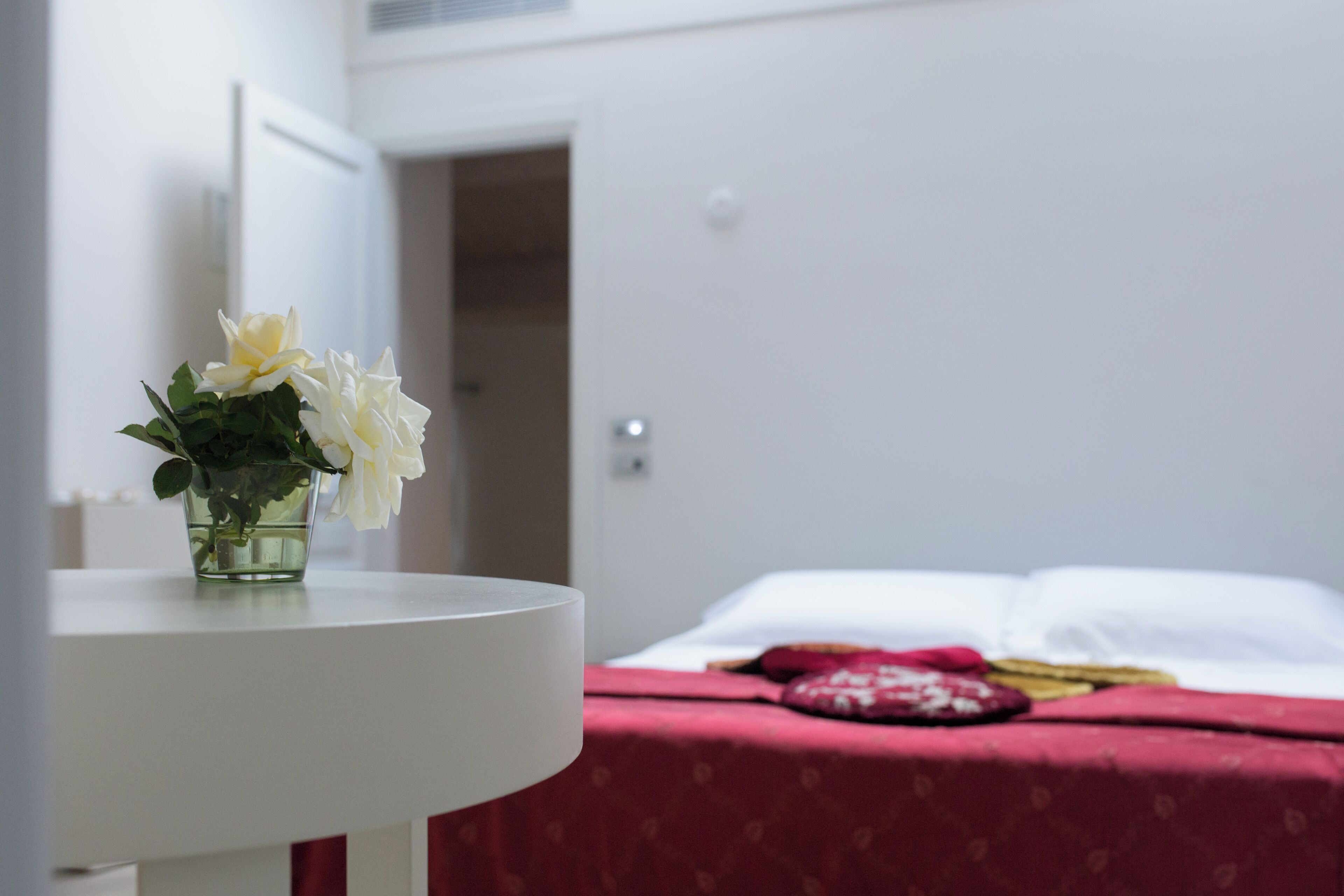 A room with a bed covered in a red blanket, a white bedside table with a vase of white flowers, and a white door in the background.