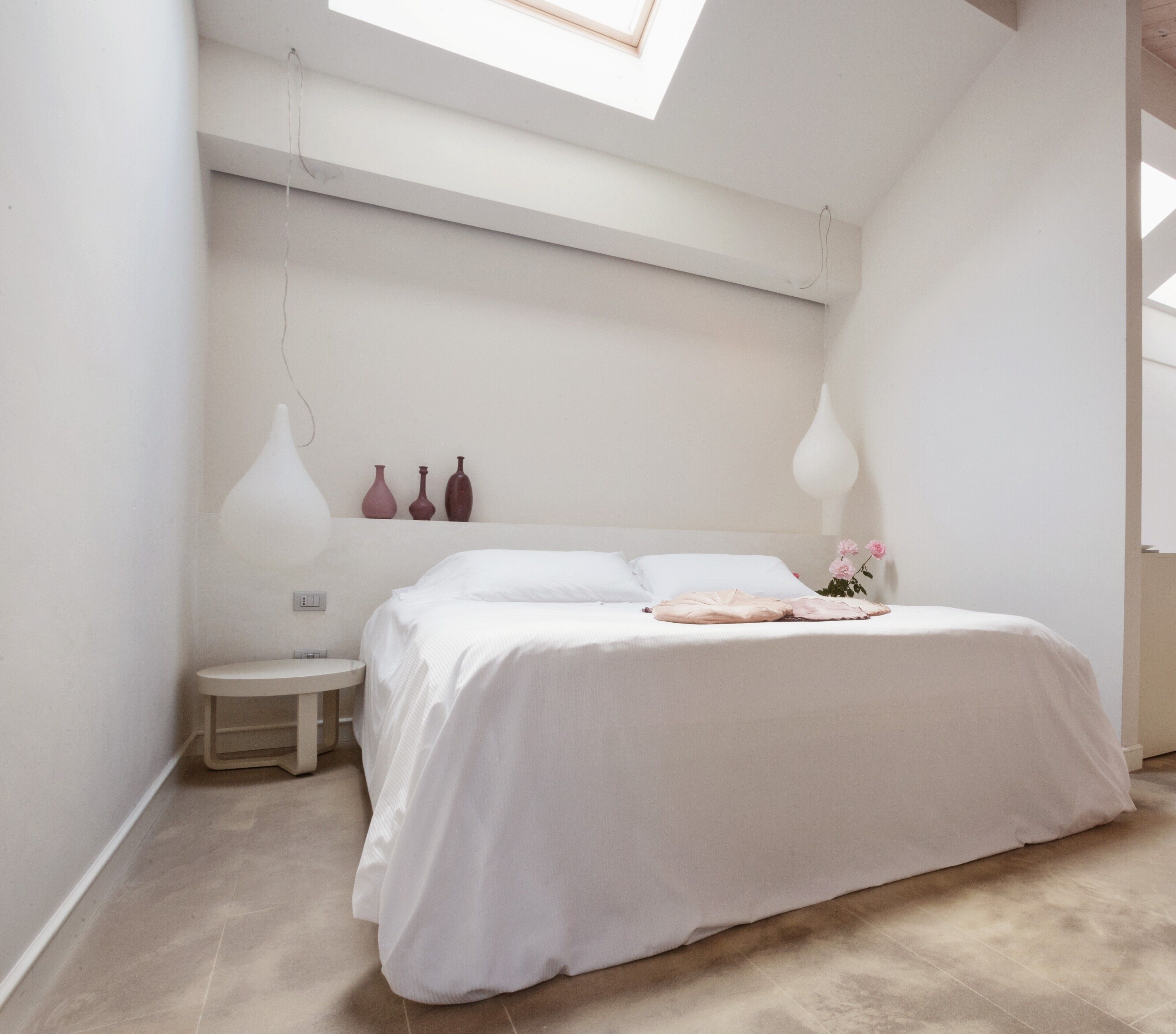 Quarto com claraboia, uma cama, mesas de cabeceira e vasos decorativos.
