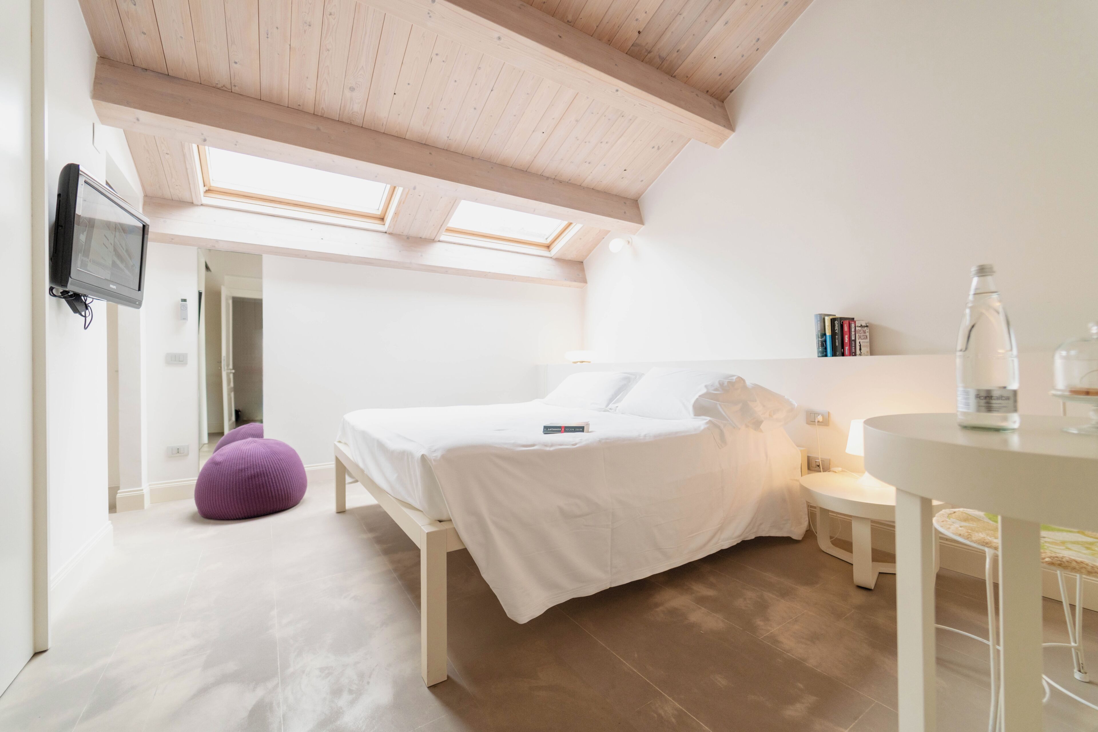 Un dormitorio con una cama, una mesita, una silla, un sillón de peluche morado, un televisor y un tragaluz.