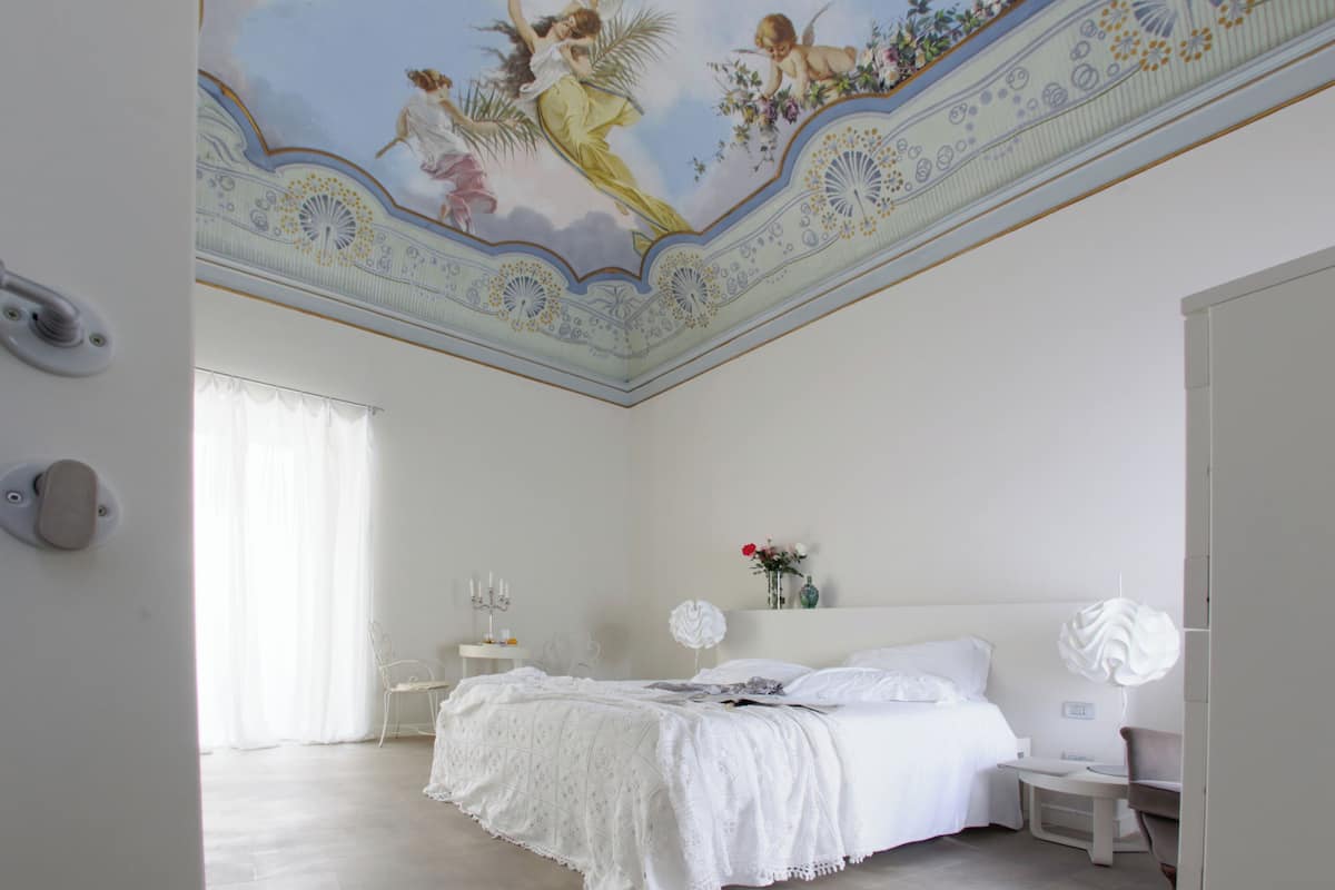 A bedroom with a decorative ceiling mural, a bed with white linens, a bedside table with a lamp, and a nightstand with a vase of flowers.