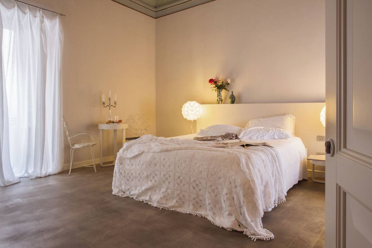 A bedroom with a large bed, a bedside table with a lamp, a chair, and a vase with flowers on the wall.