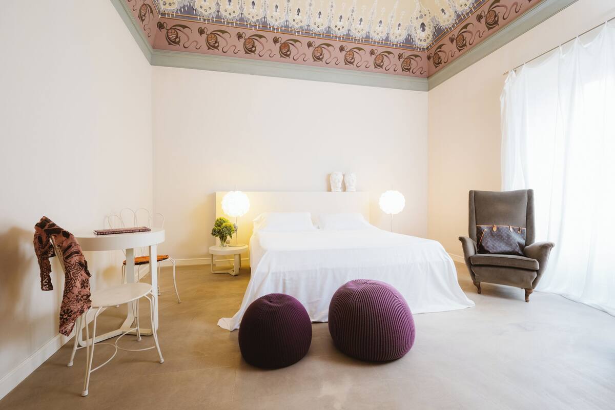 A hotel room with a bed, a chair, a table, and two ottomans.