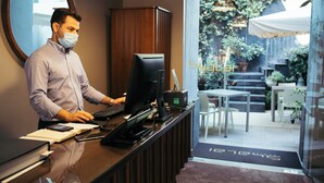 Un comptoir d’accueil avec un ordinateur, un téléphone et un bloc-notes. Un miroir est accroché au mur, et une porte donne accès à un espace extérieur aménagé avec des tables et des chaises.