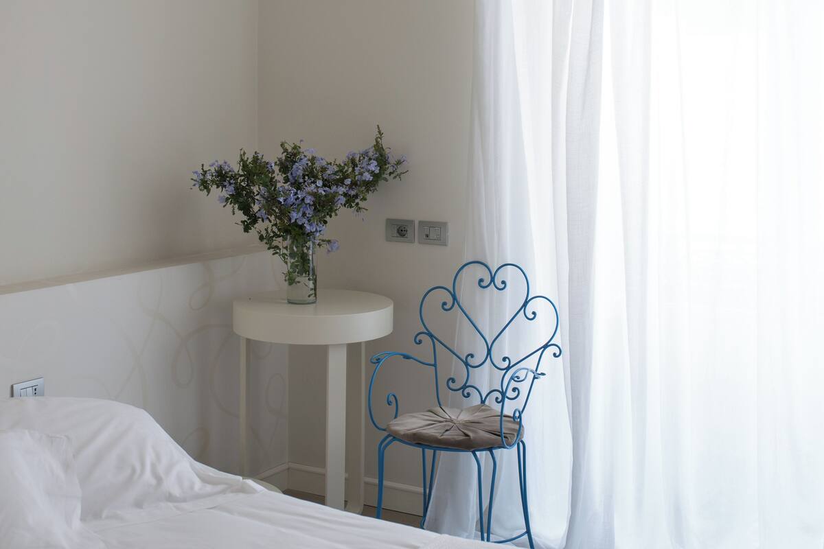 A bed, a small round table with a vase of flowers, and a blue chair in a room with white walls and a white curtain.