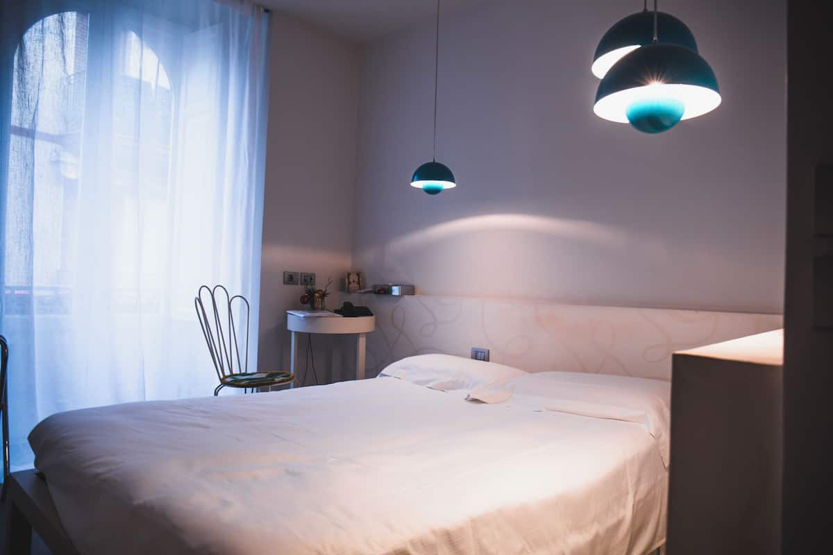 A neatly made bed with white linens, a bedside table with a lamp, and a chair by the window.