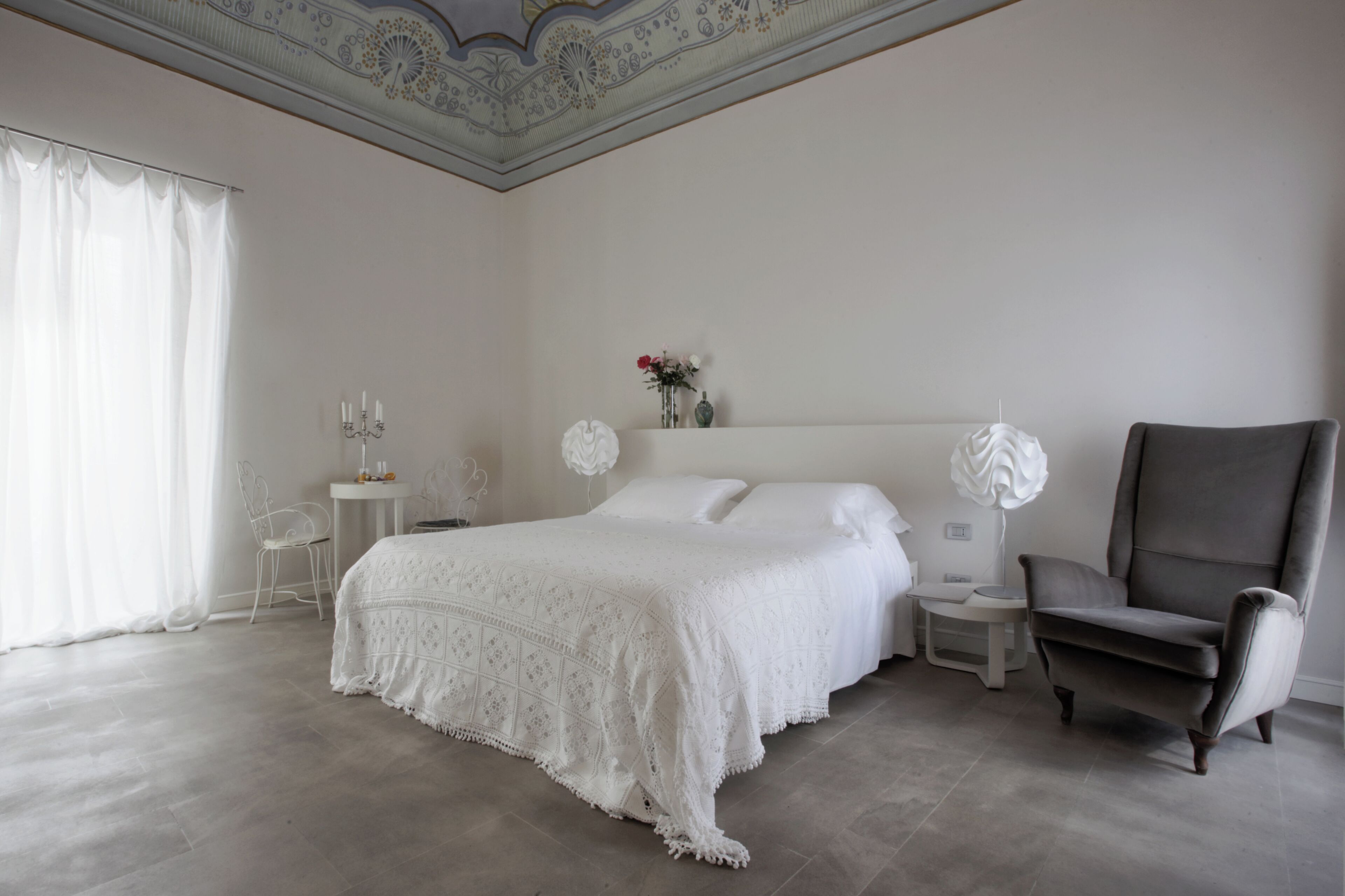 Un dormitorio con una cama grande, una silla, una mesita de noche y un jarrón con flores.