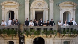 Un groupe de personnes se trouve sur un balcon orné de balustrades décoratives, et la façade de l’immeuble présente une architecture de style classique.