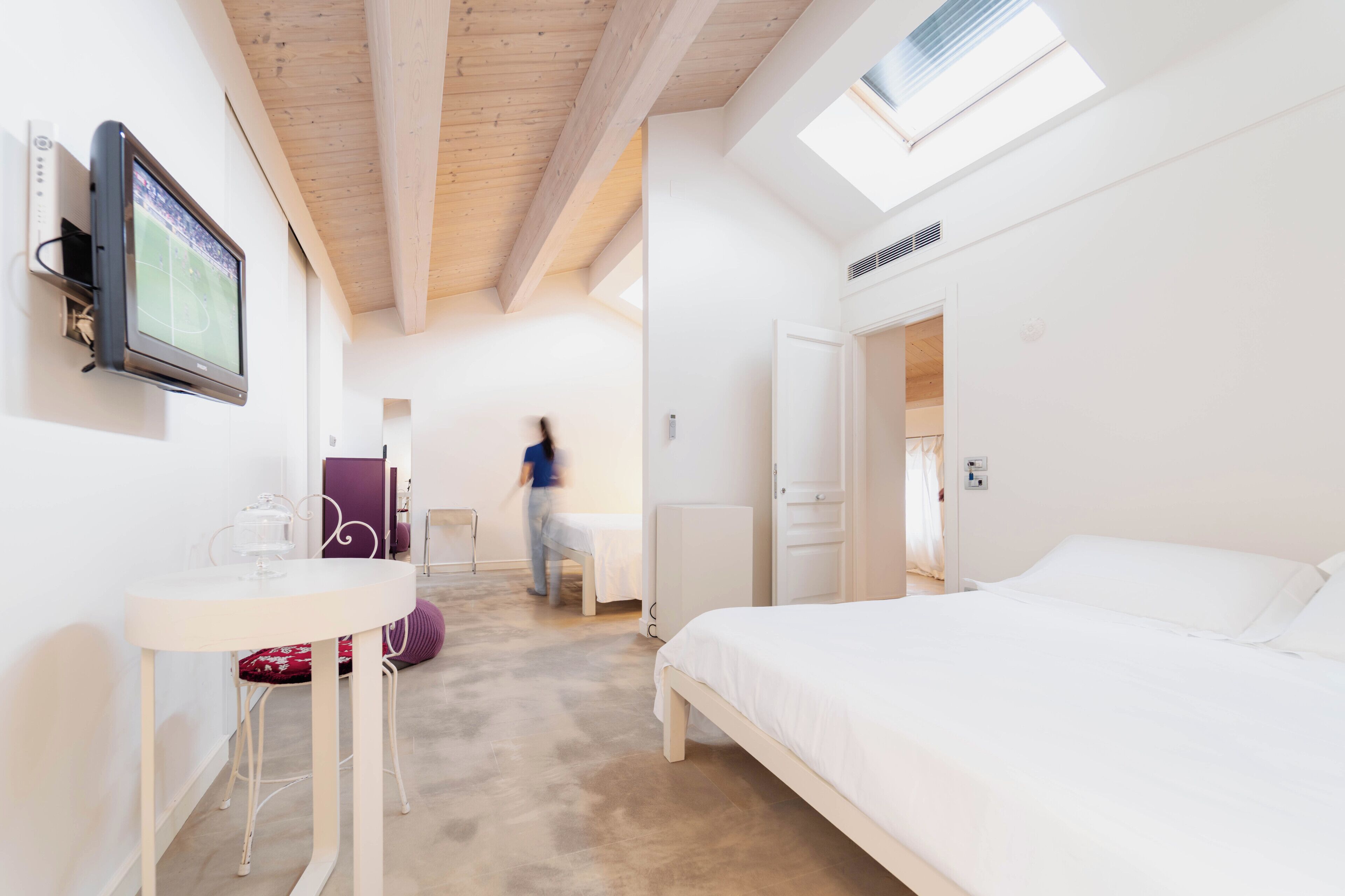 A bedroom with a bed, a TV mounted on the wall, a table with a chair, and a door leading to another room.