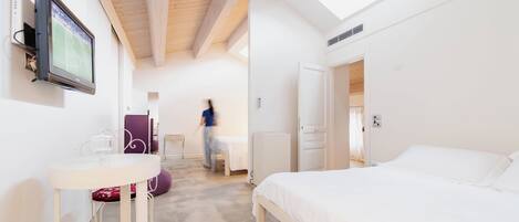A bedroom with a bed, a TV mounted on the wall, a table with a chair, and a door leading to another room.