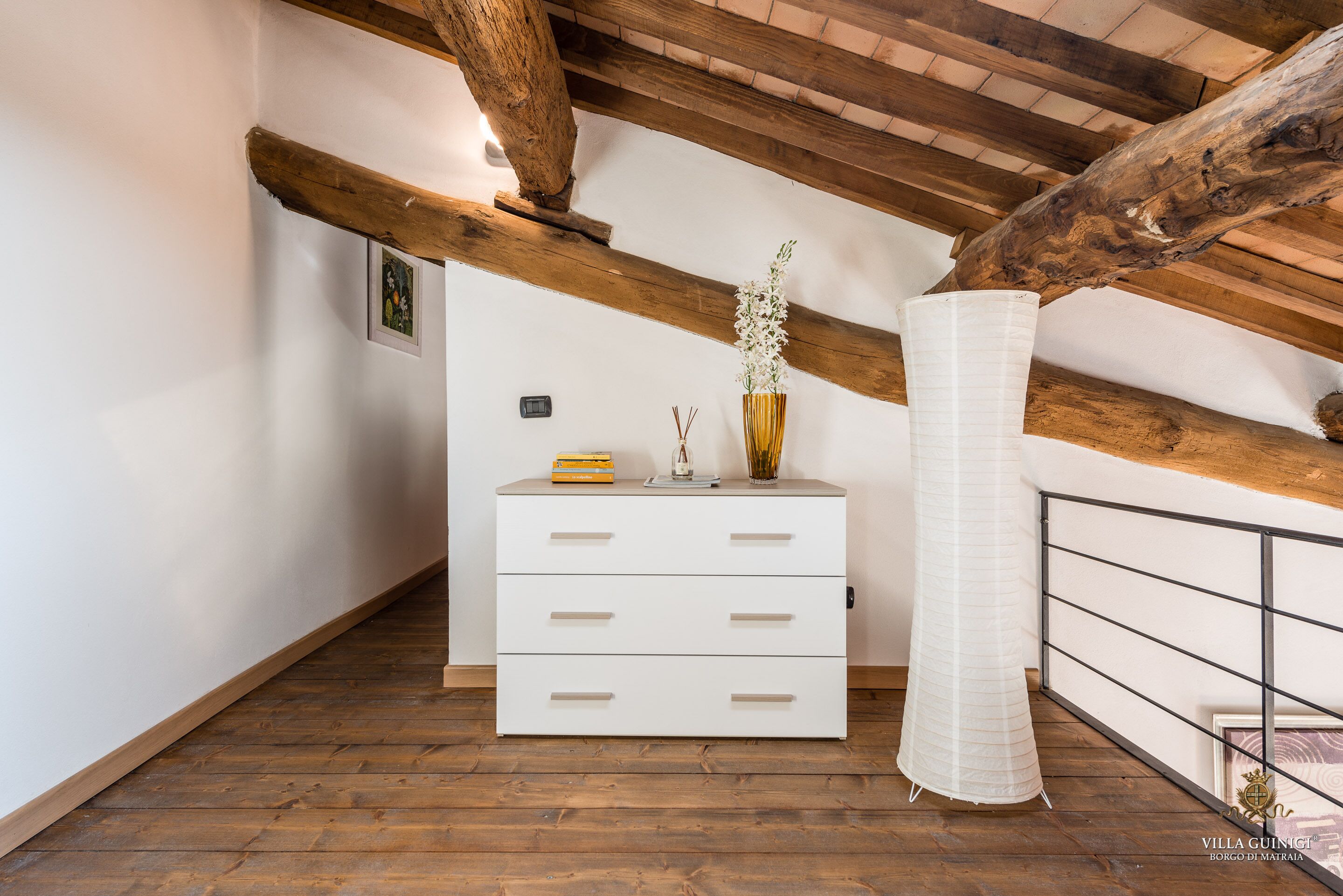 A white dresser with drawers, a vase with flowers, and a wooden ceiling beam in a room with wooden flooring and a white wall.
