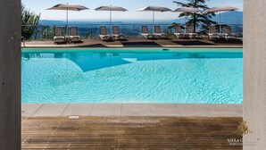 A pool area with lounge chairs and umbrellas, a wooden deck, and a clear view of the ocean and mountains.