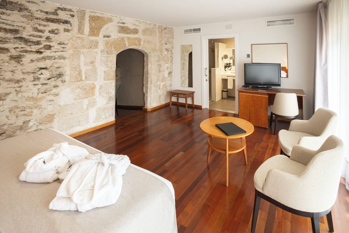 A hotel room with a stone wall, wooden flooring, a sofa, a small table, a TV, and a chair.