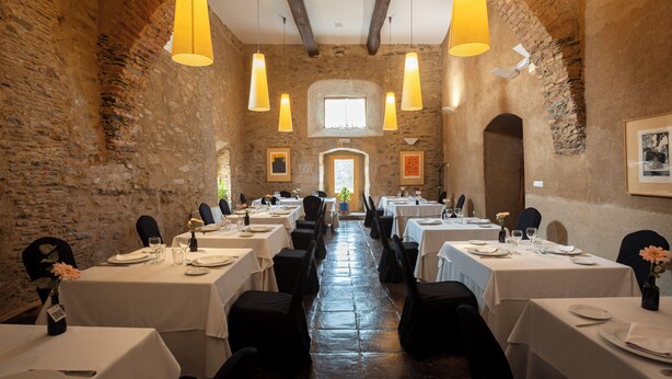 A long dining hall with tables set for a meal, featuring stone walls and hanging pendant lights.