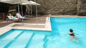A swimming pool with lounge chairs and umbrellas, a stone wall in the background, and a person swimming.