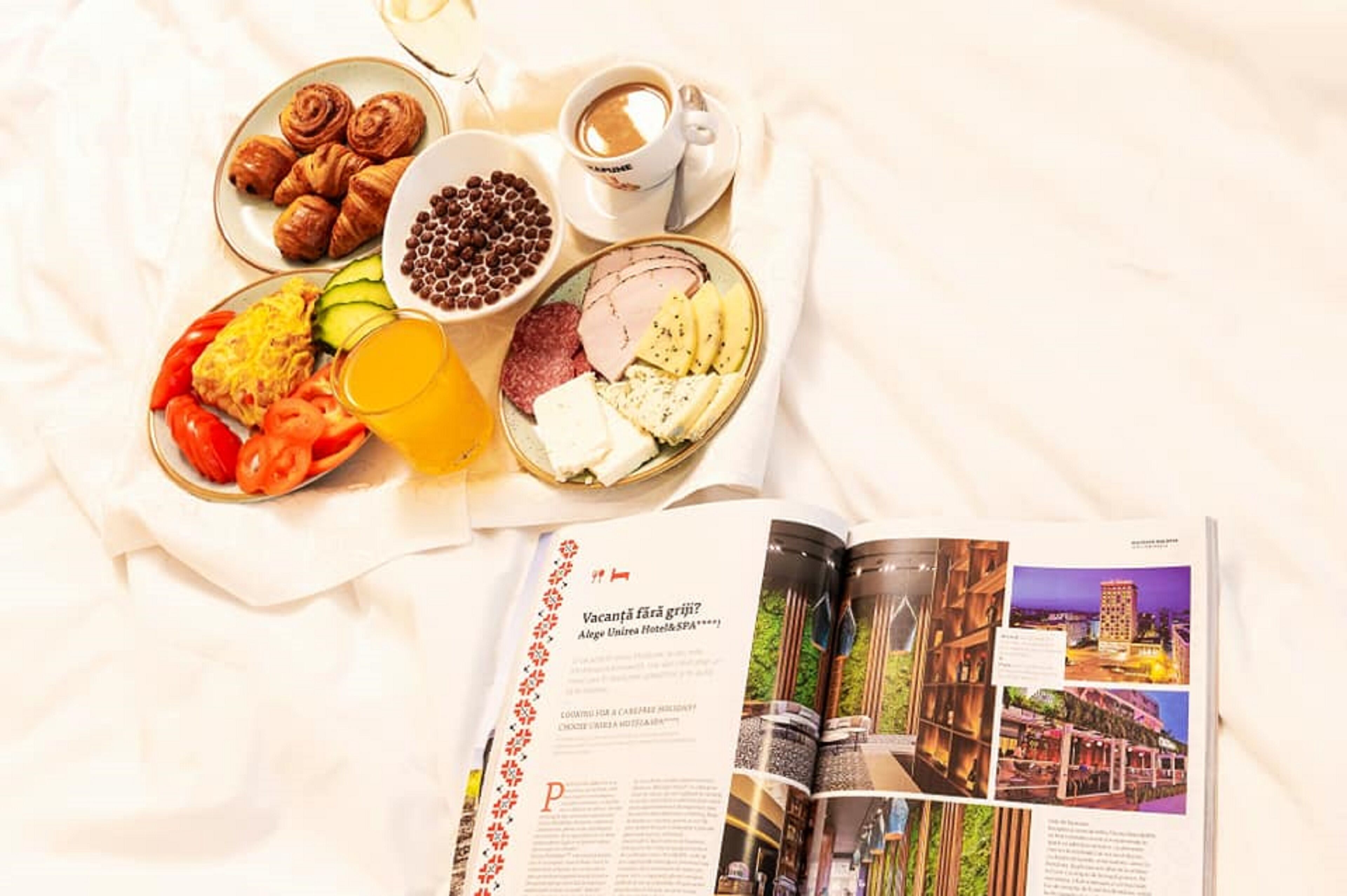 A breakfast spread with croissants, coffee, and various fruits and cheeses on a bed, with a magazine open beside it.