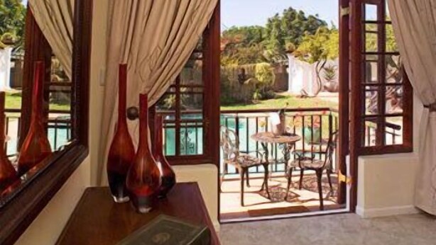 A room with a view of a garden through a glass door, a wooden table with a book and vases, and curtains on the windows. - Three Palms Luxury Cottage (Cape Town)