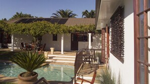 A pool area with a wooden deck, a potted plant, and a building with a tiled roof and white walls. - Three Palms Luxury Cottage (Cape Town)