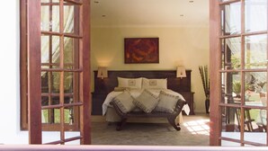 A bedroom with a large bed, two bedside tables, a painting on the wall, and a view of the outdoors through a window. - Three Palms Luxury Cottage (Cape Town)