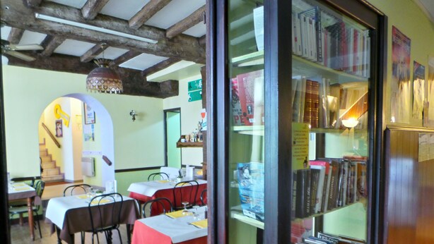 A dining area with tables and chairs, a staircase, and a bookshelf filled with books. - Hôtel La Fontaine (Lourdes)