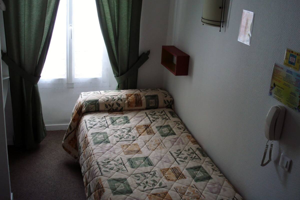 A small, neatly made bed with a patterned bedspread, a red bedside table, a wall-mounted telephone, and a window with green curtains.