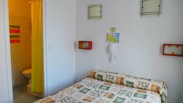 Un dormitorio con una cama, un baño visible a través de una puerta abierta y un teléfono instalado en la pared.