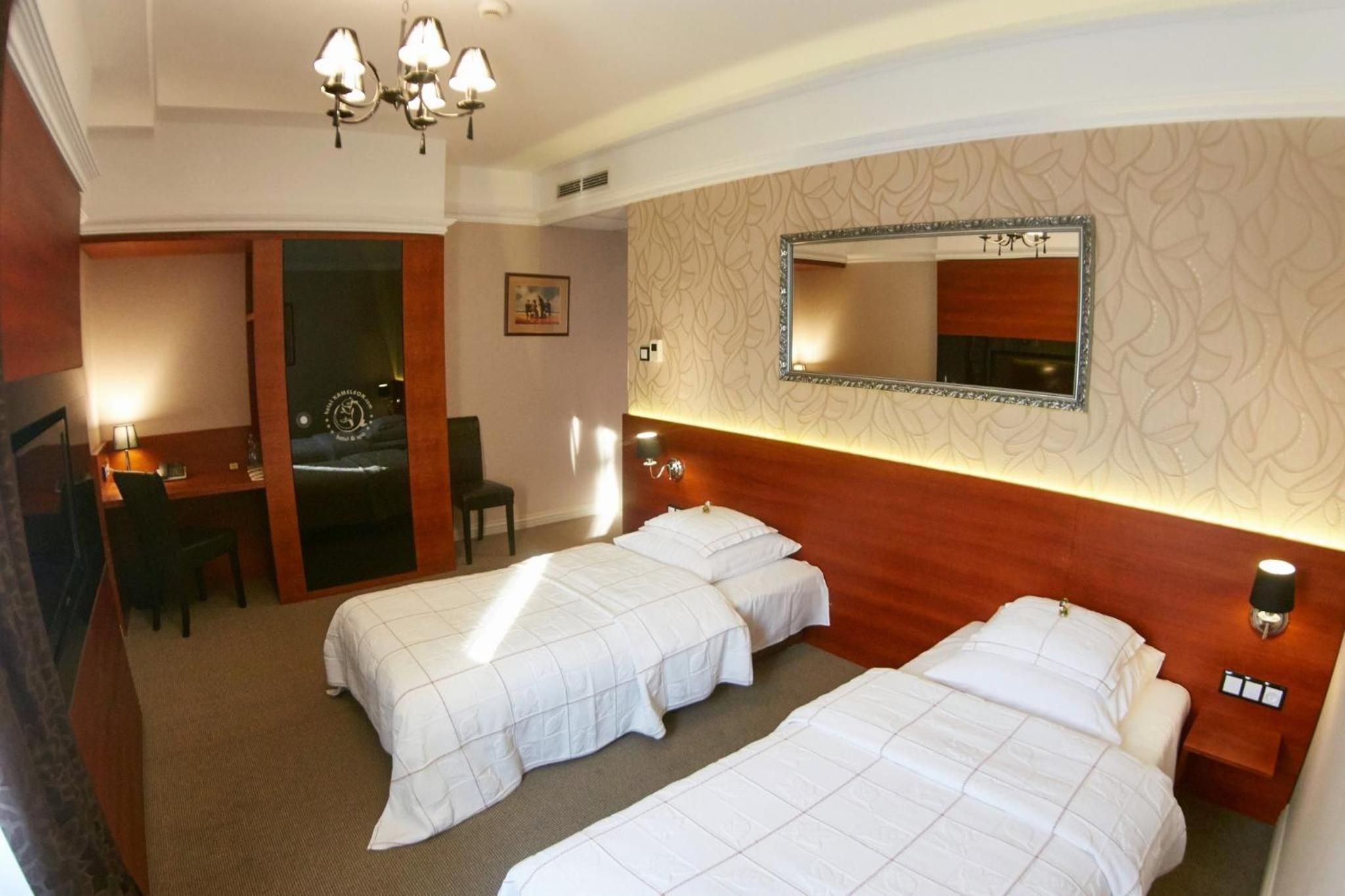 A hotel room with two beds, a desk, a chair, a mirror, and a chandelier.