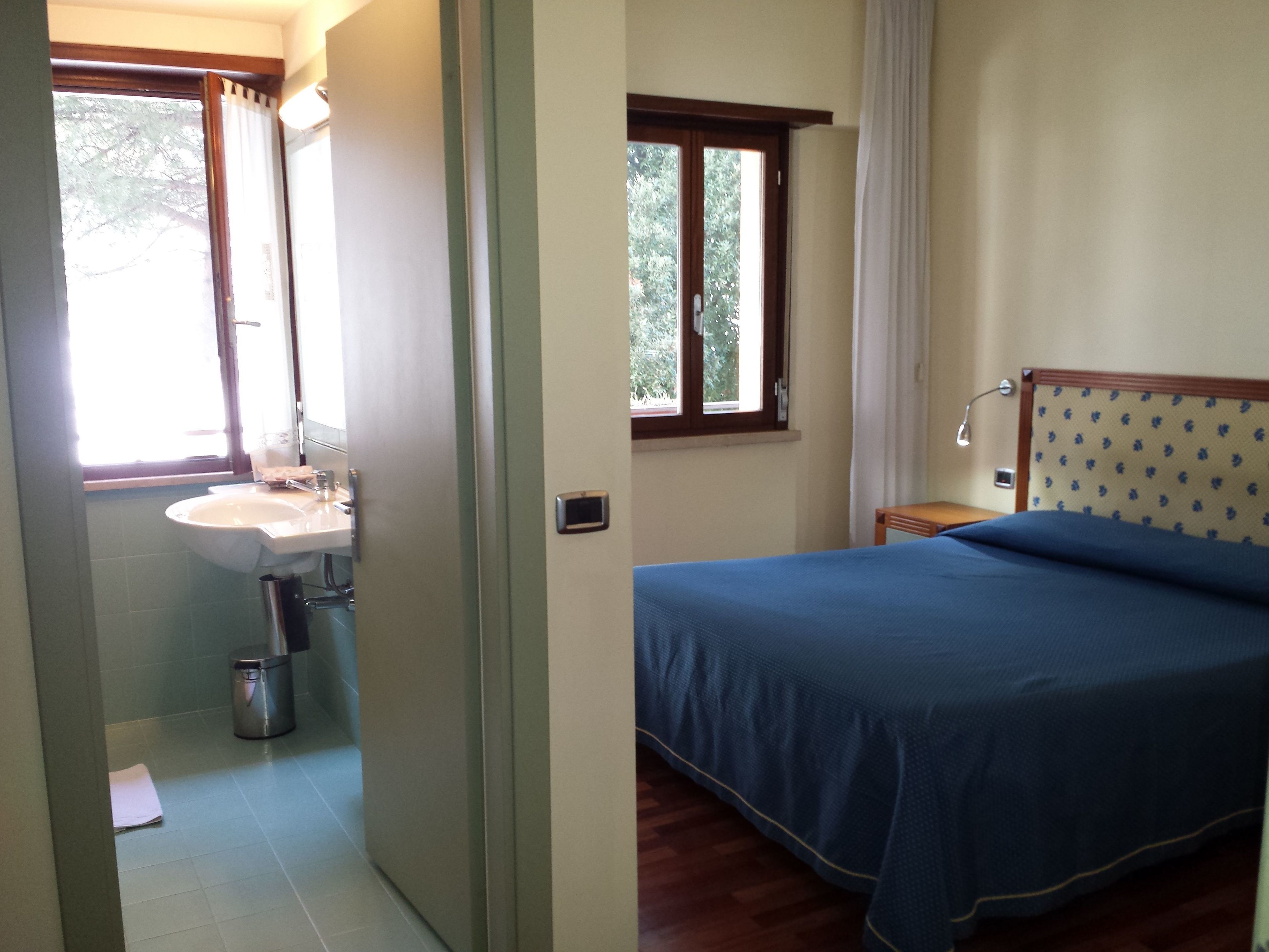 Ein Schlafzimmer mit Bett, Nachttisch, Fenster und eigenem Badezimmer mit Waschbecken und Spiegel.