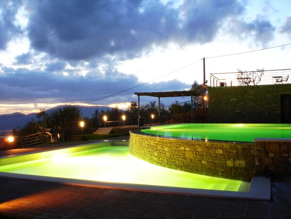 A swimming pool with a stone wall, surrounded by a covered seating area and a view of mountains.