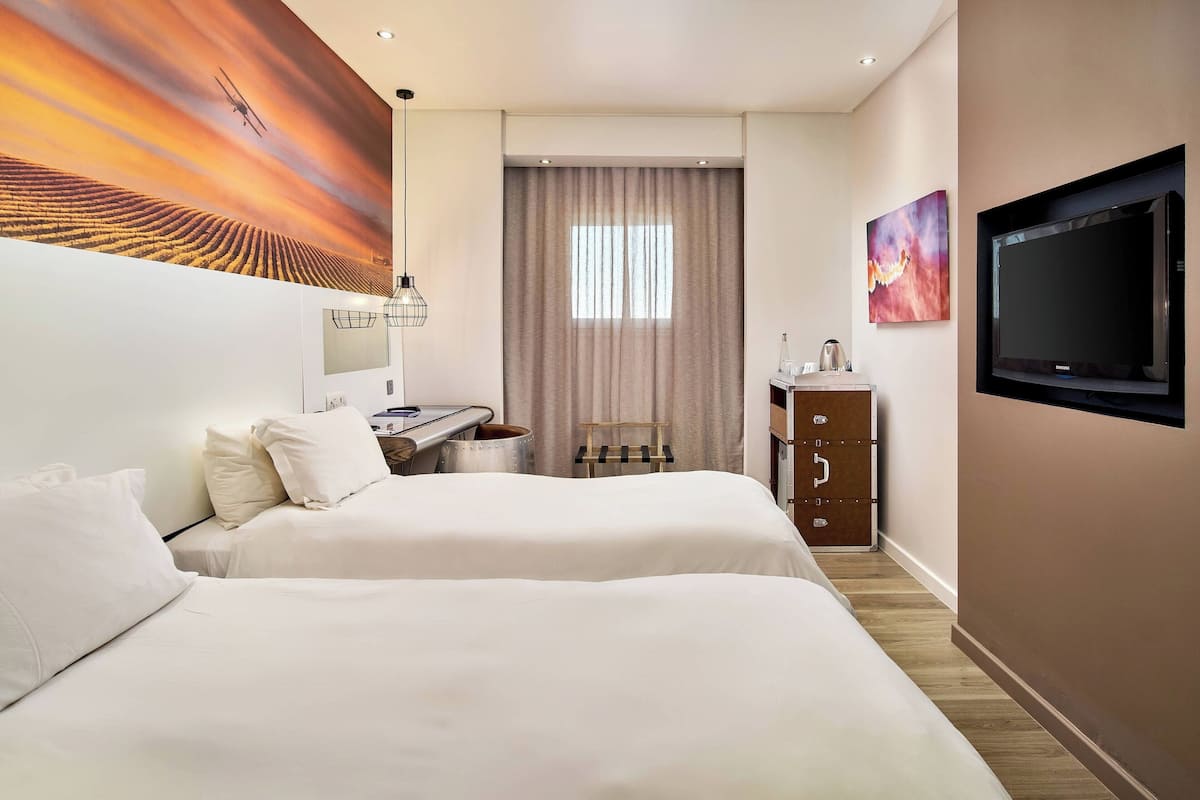 Ein Hotelzimmer mit zwei Betten, einem Schreibtisch mit Stuhl, einem Fernseher, einem Bild und einem Fenster mit Vorhängen.