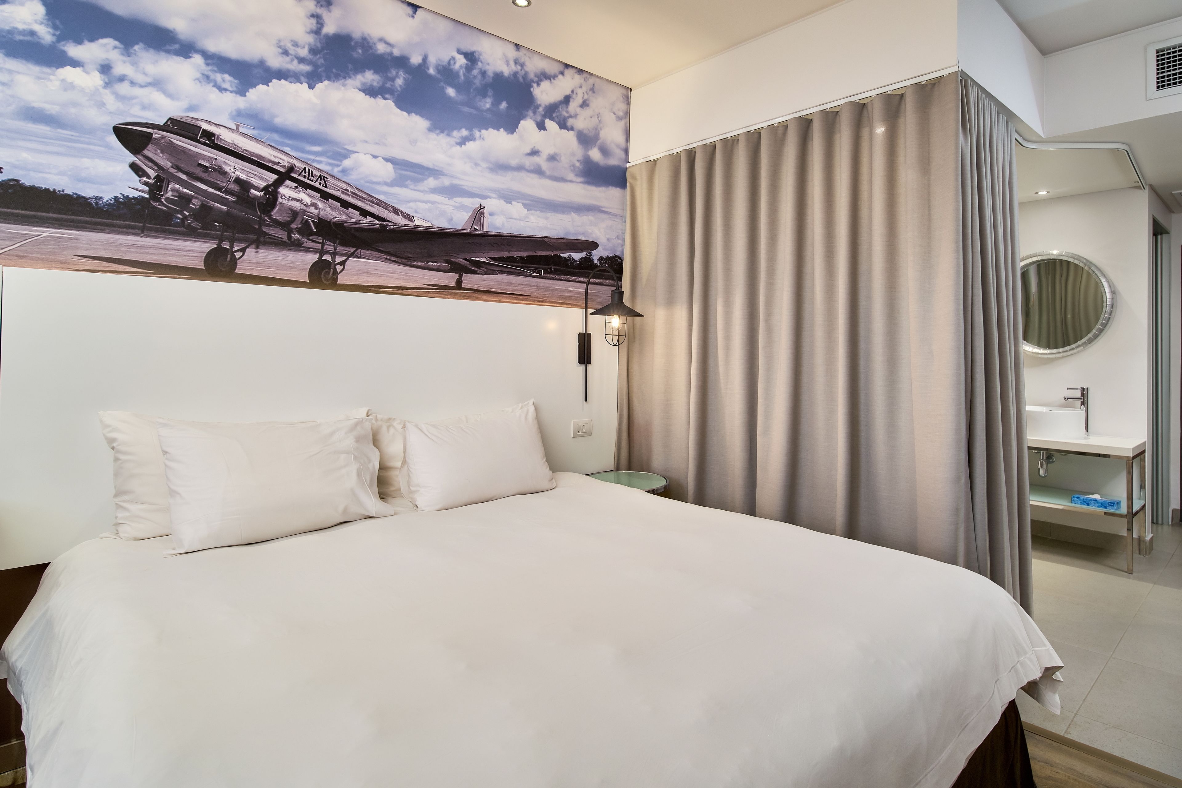 Habitación de hotel con una cama grande, una mesita de noche, una lámpara, un baño con espejo y un mural de un avión en la pared.