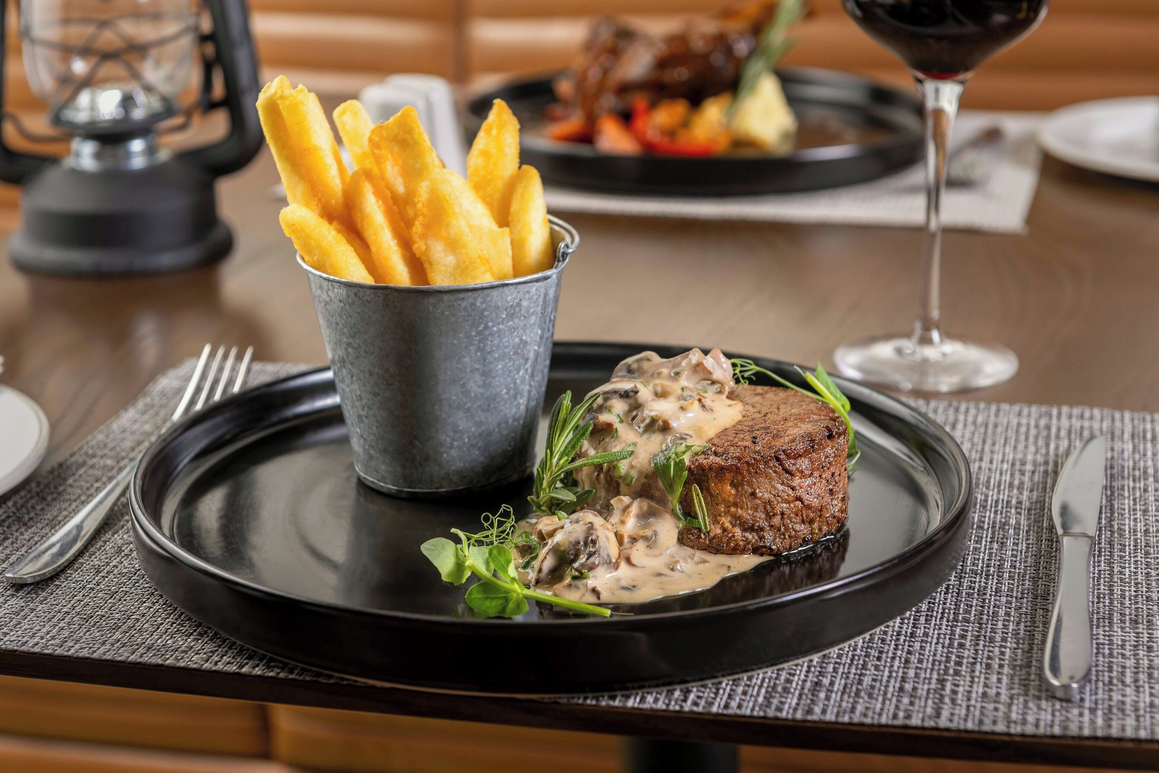 Een bord met steak en champignons, een emmer friet en een glas rode wijn op een tafel.