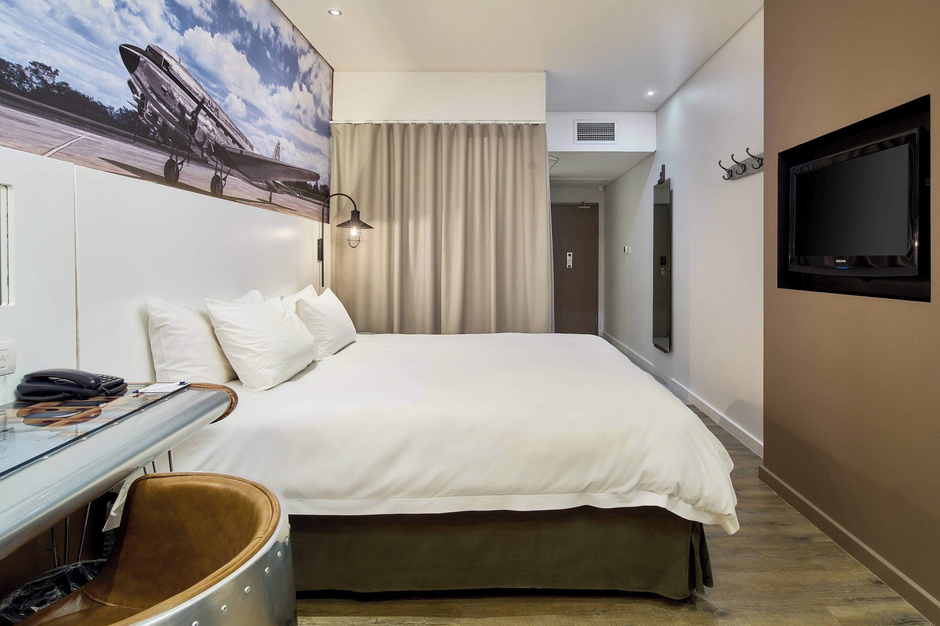 A hotel room with a bed, a desk with a chair, a television, and a large window with curtains.