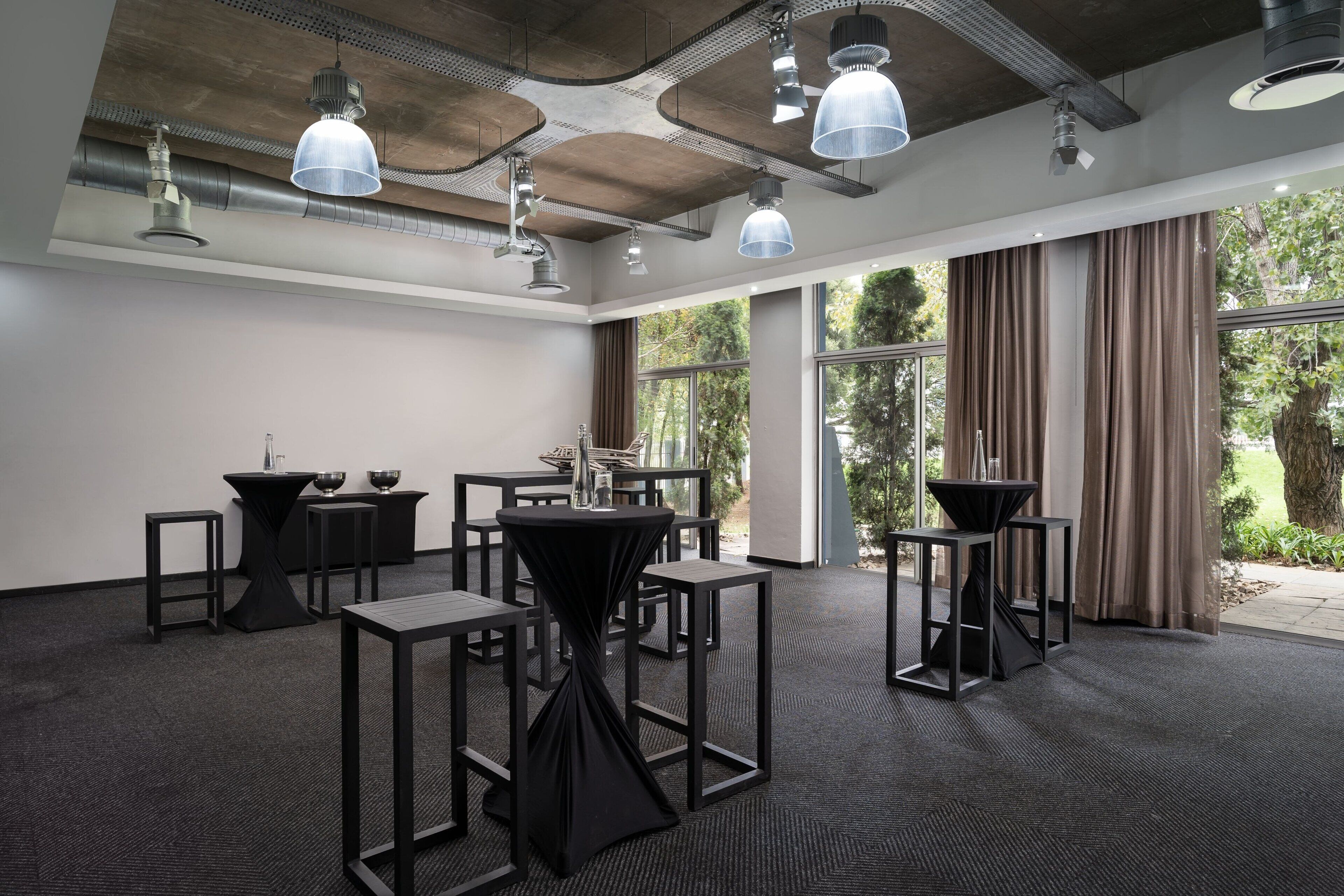 A conference room with multiple tables and chairs, large windows with curtains, and industrial-style ceiling lighting.