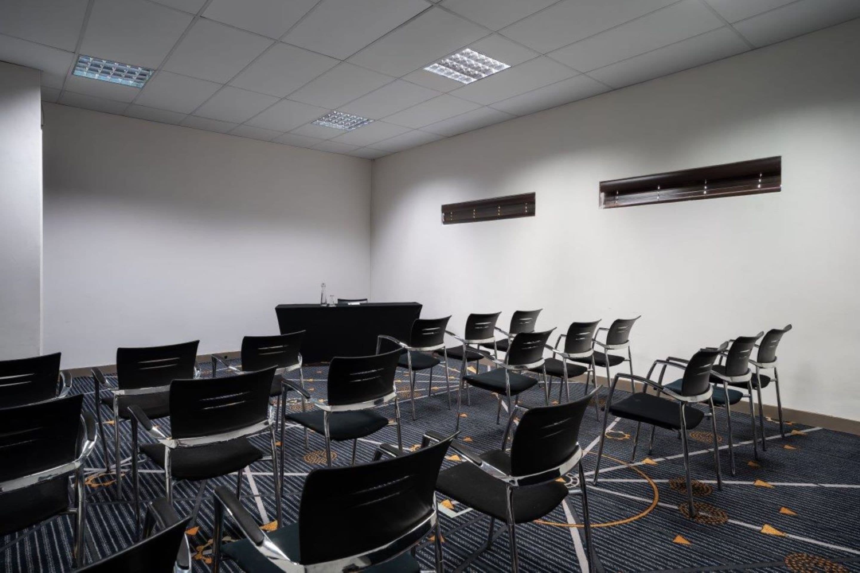 Uma sala de conferências com fileiras de cadeiras pretas, um tapete estampado e um teto branco com iluminação embutida.