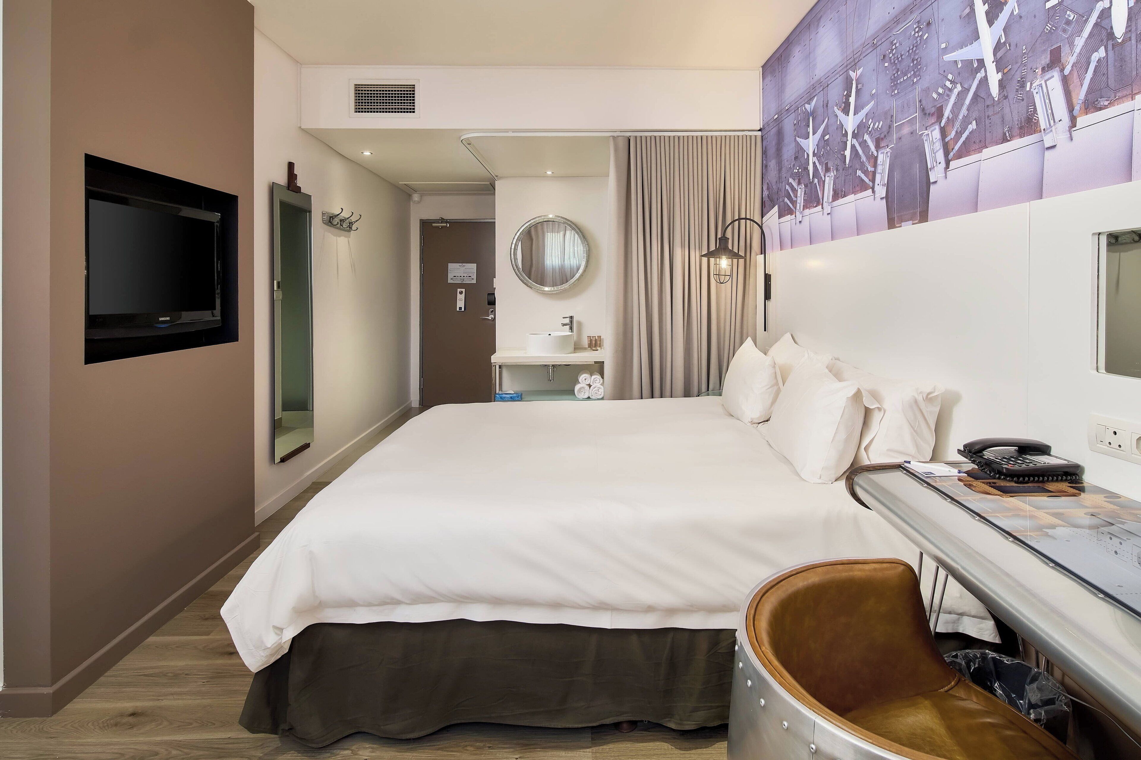 A hotel room with a large bed, a desk with a telephone, a mirror, and a wall-mounted TV.