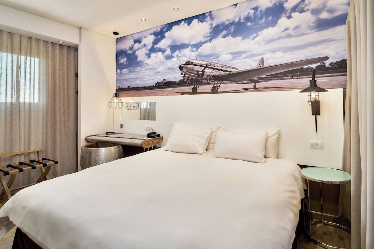 Ein Hotelzimmer mit einem großen Bett, einem Schreibtisch mit Stuhl, einem kleinen Tisch, einer Lampe und einem großen Fenster mit Vorhängen.