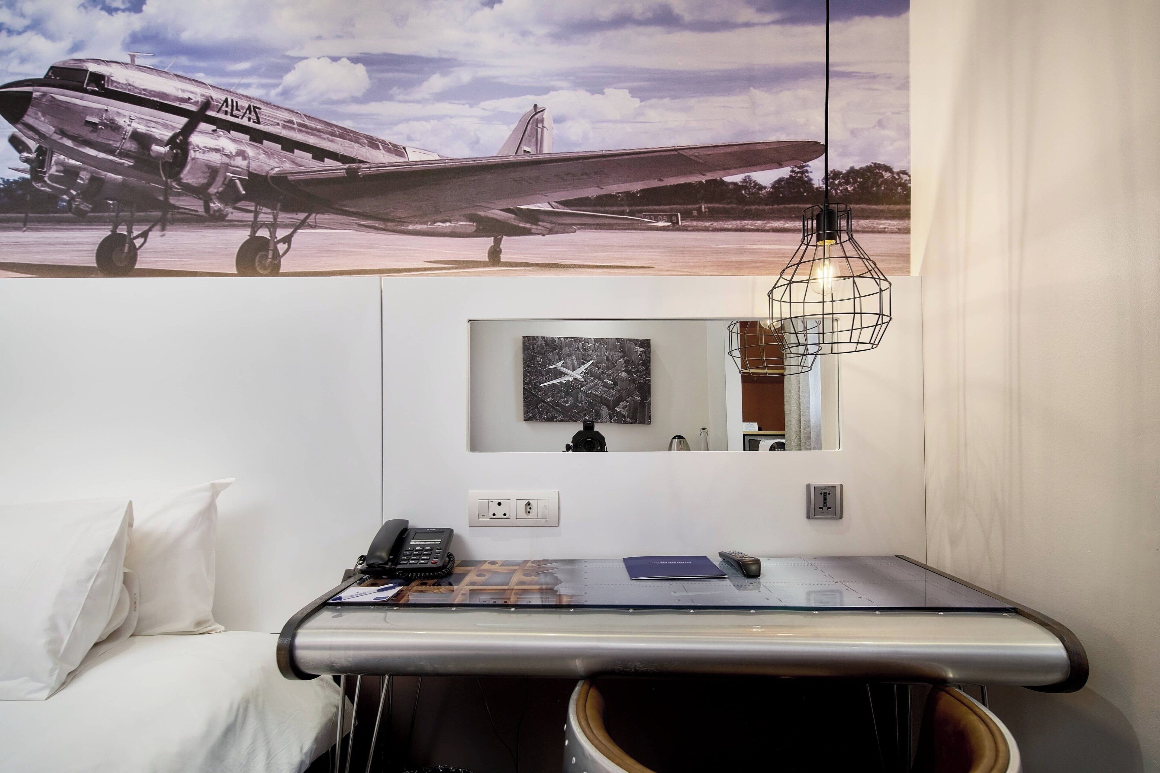 A hotel room with a large airplane poster on the wall, a desk with a telephone, a clock, and a hanging light fixture.