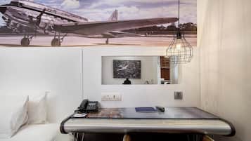 A hotel room with a large airplane poster on the wall, a desk with a telephone, a clock, and a hanging light fixture.