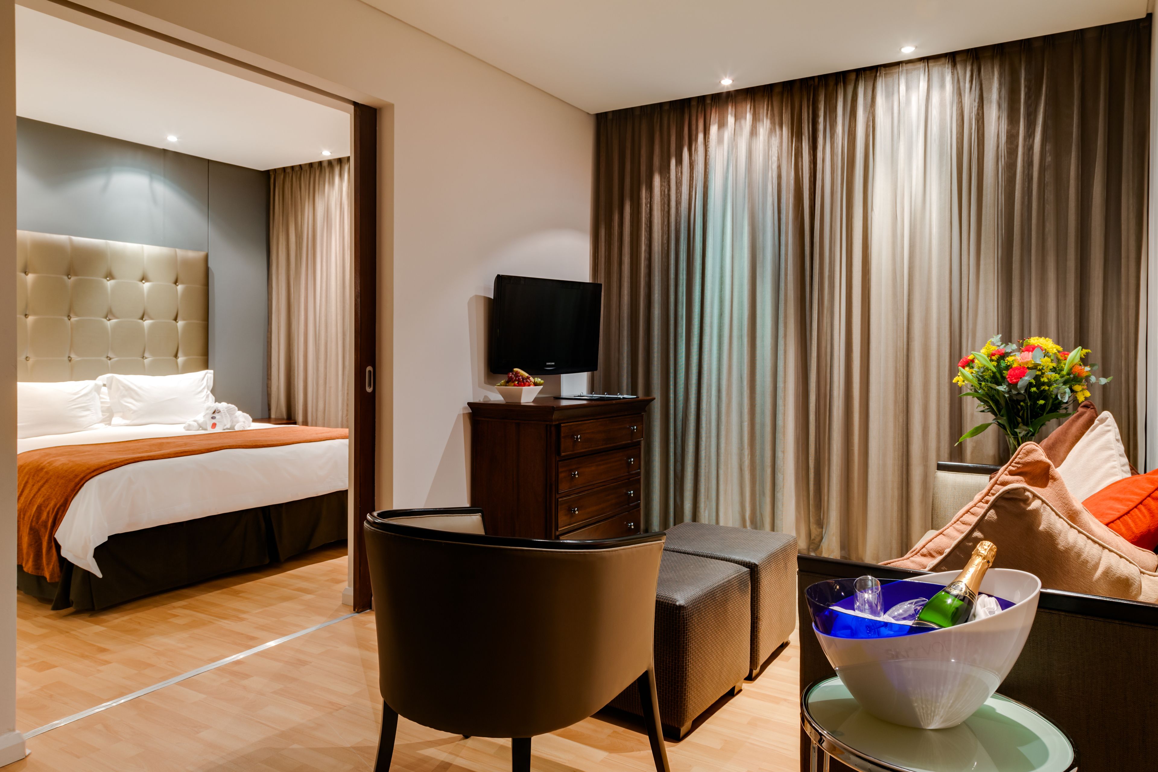 Une chambre d’hôtel comprenant un lit, une télévision, une commode, une chaise, une table avec une coupe de champagne et un canapé agrémenté d’un arrangement floral.