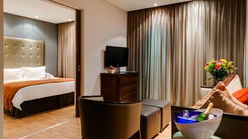 A hotel room with a bed, a TV, a dresser, a chair, a table with a bowl of champagne, and a sofa with a flower arrangement.