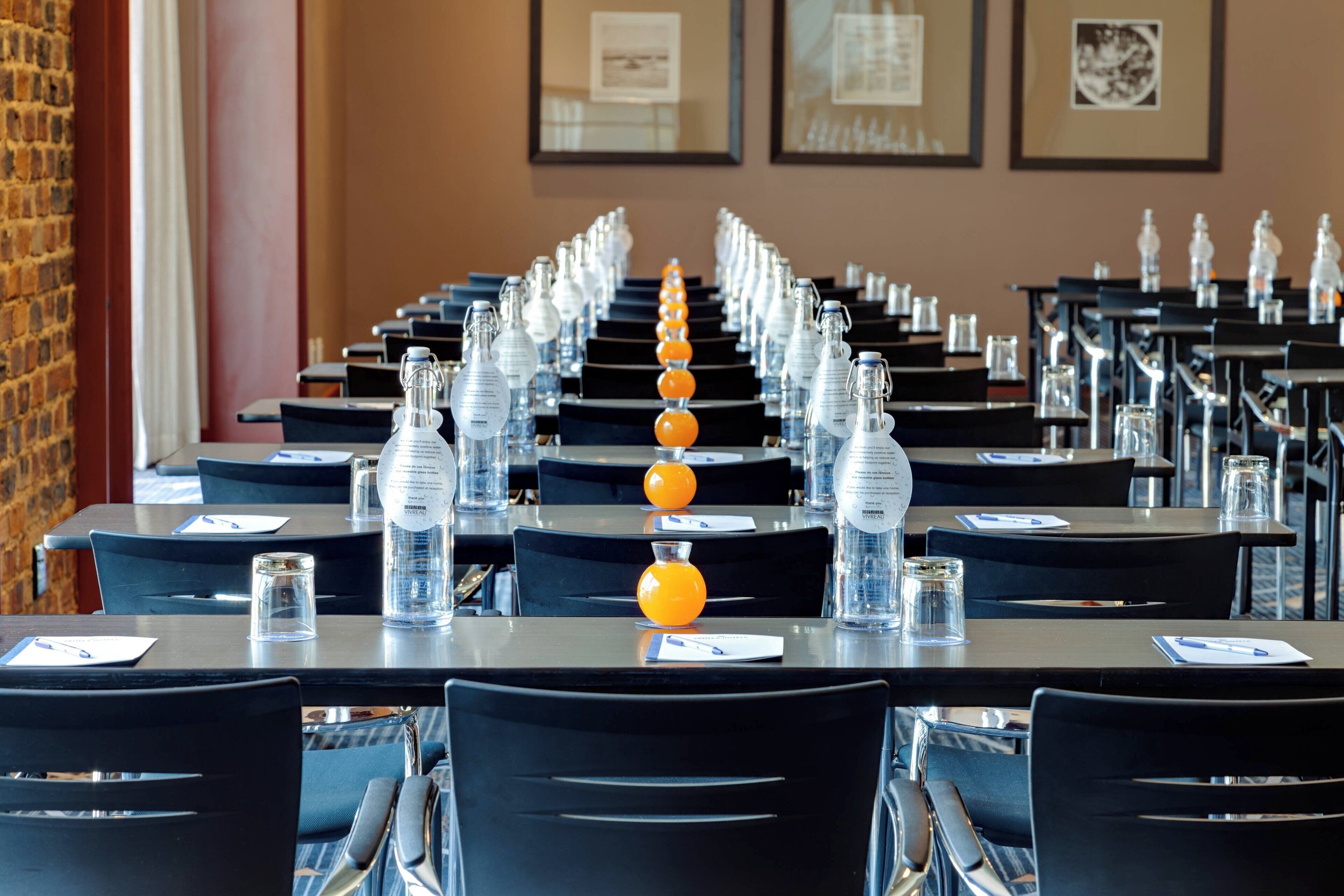 Un salón de conferencias con filas de mesas, cada una con una botella de vidrio transparente y un pequeño objeto naranja.
