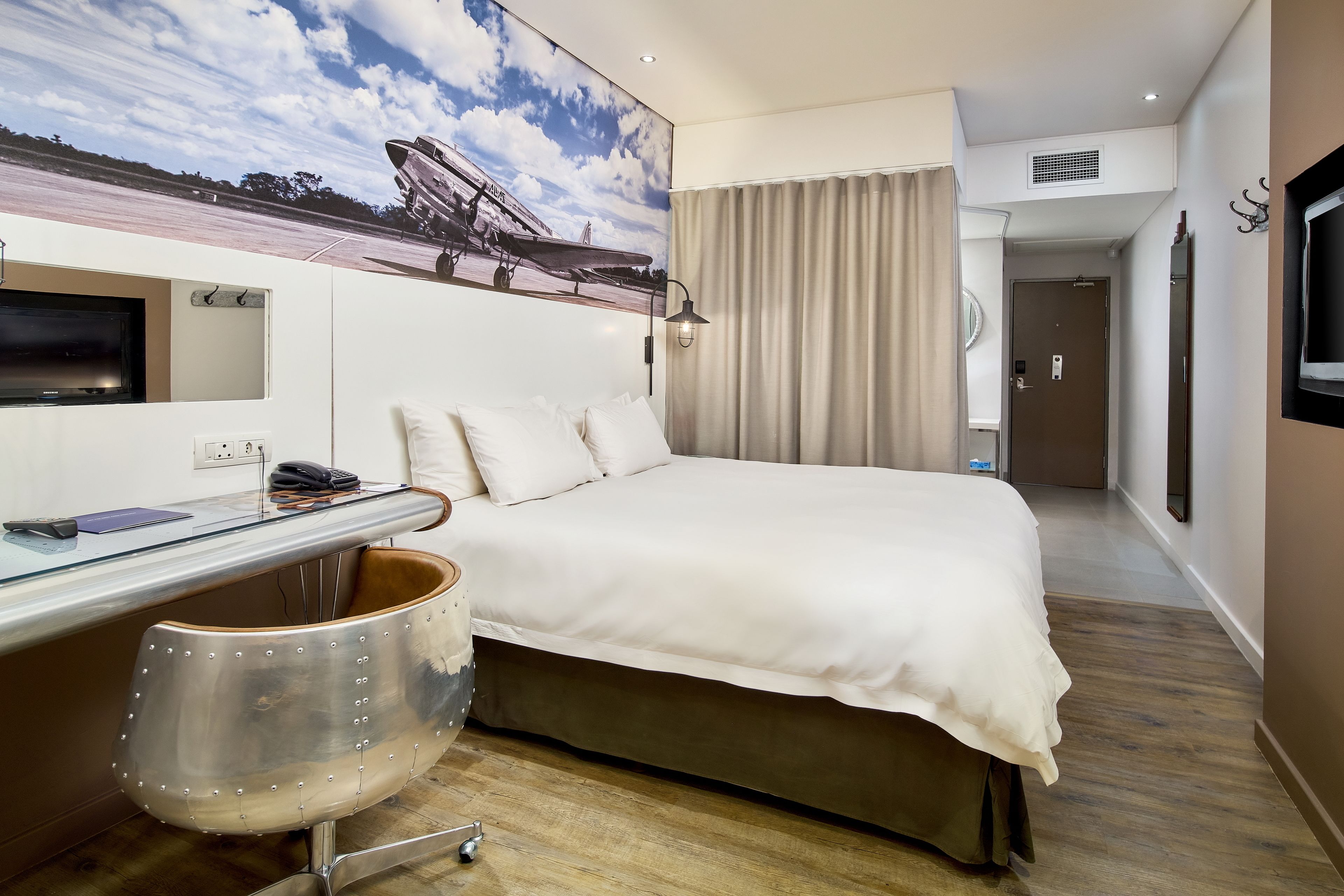 Habitación de hotel con cama, escritorio, silla, televisor y un cuadro de un avión en la pared.