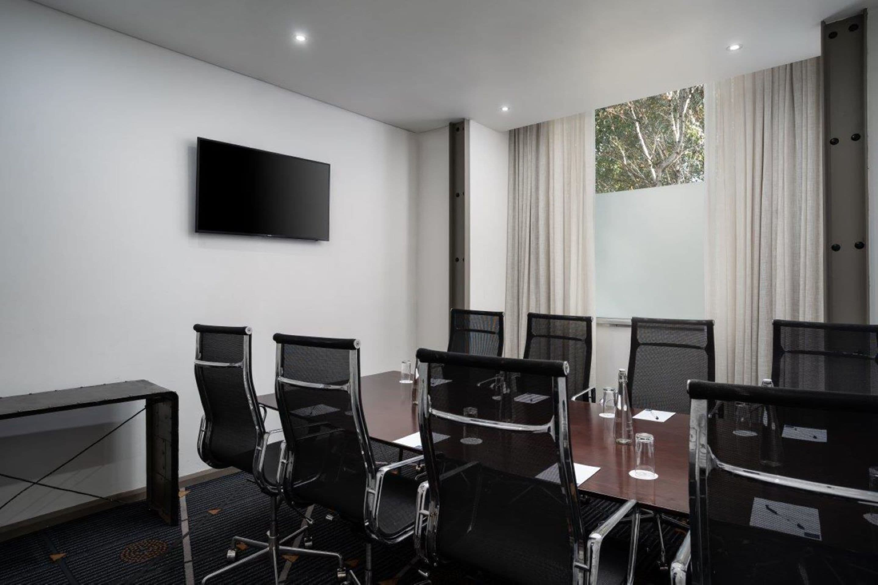 A conference room with a large table, six chairs, a flat-screen TV, and a window with curtains.