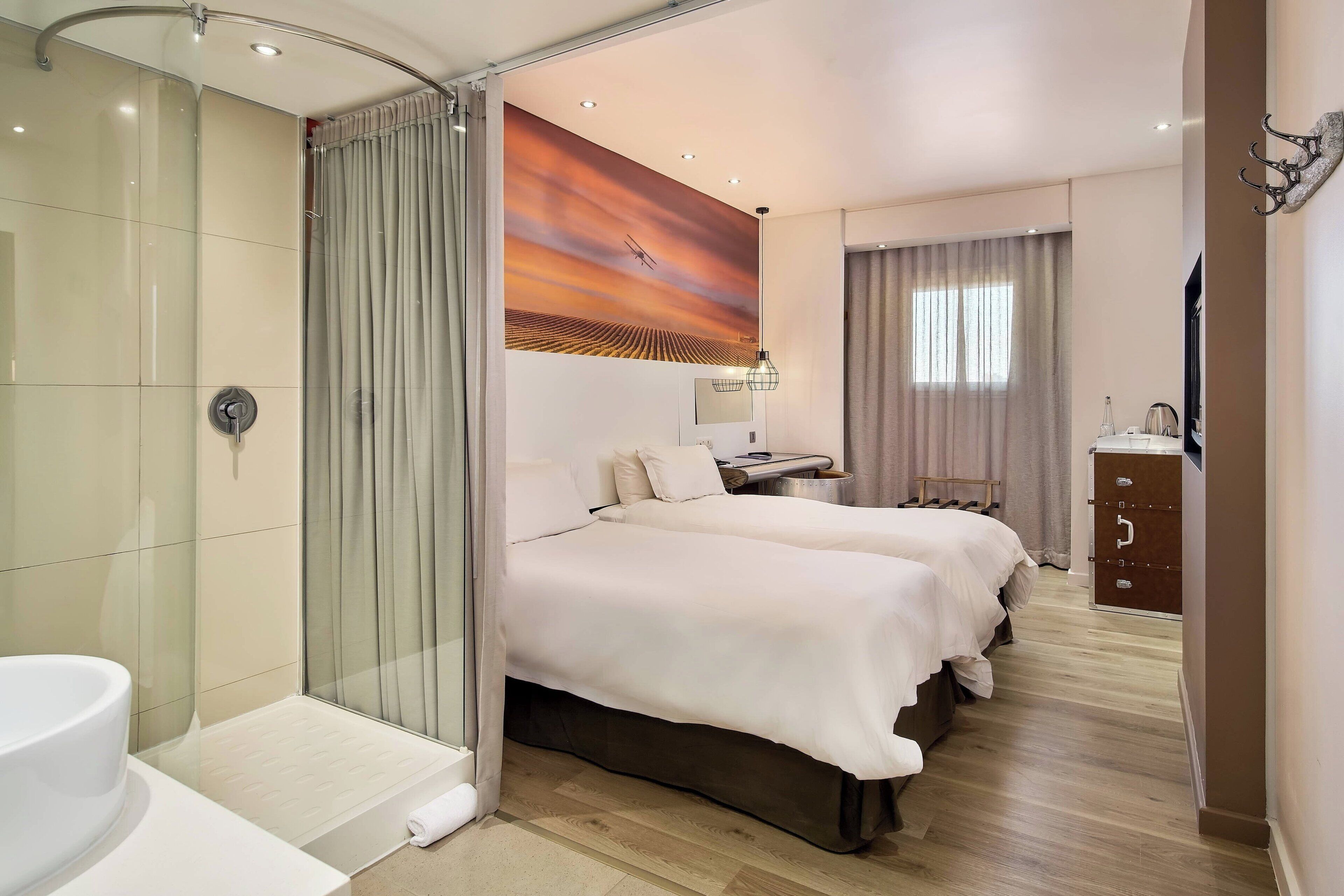 A hotel room with a glass-enclosed shower, two beds with white bedding, a painting on the wall, a desk with a lamp, and a wardrobe.