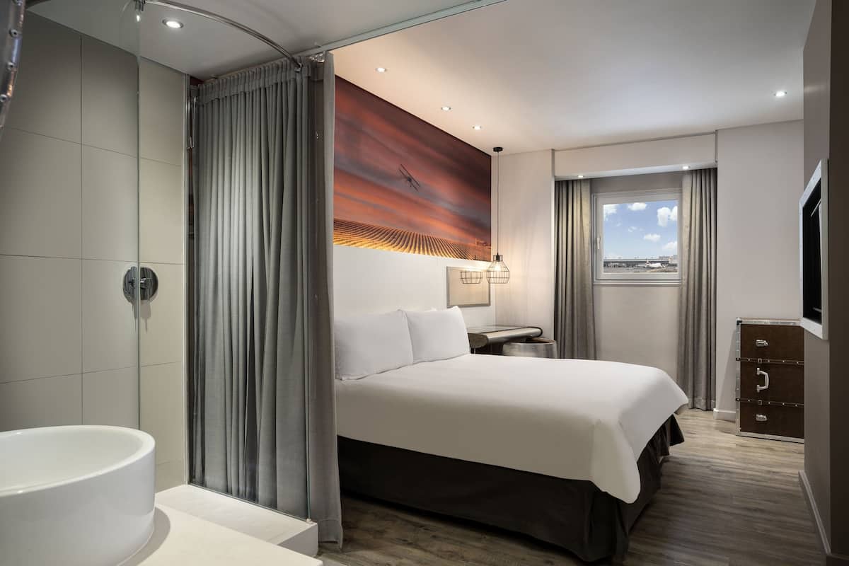 Ein modernes Hotelzimmer mit einem großen Bett, einem Badezimmer mit Badewanne, einem Schreibtisch mit Lampe und einem Fenster mit Blick auf die Stadt.